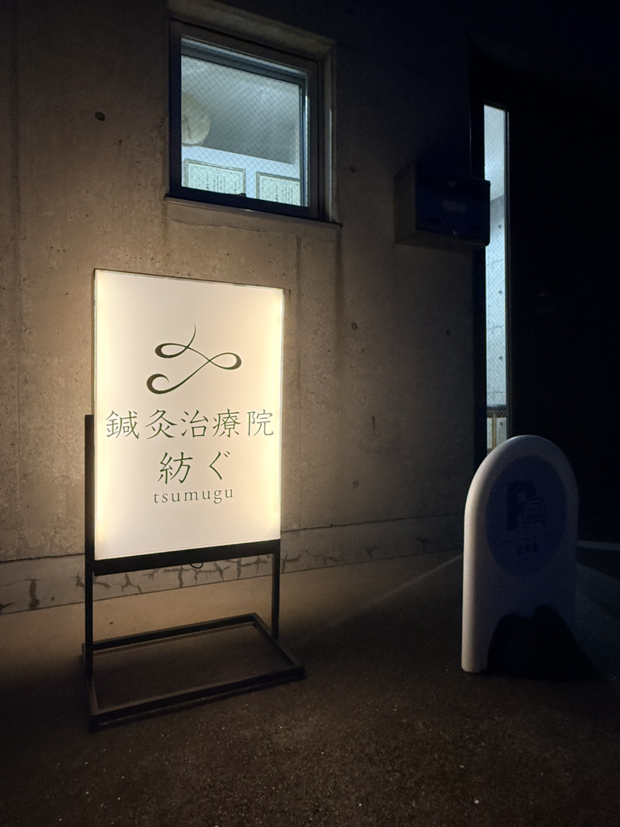 Illuminated sign for a Japanese acupuncture clinic named "tsumugu" outside a concrete building at night