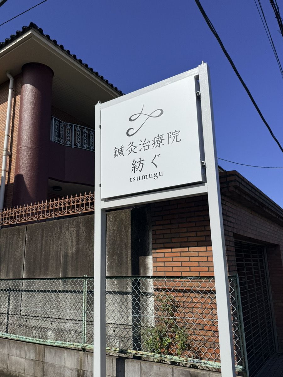 White signage for "tsumugu" acupuncture clinic beside brick building against blue sky